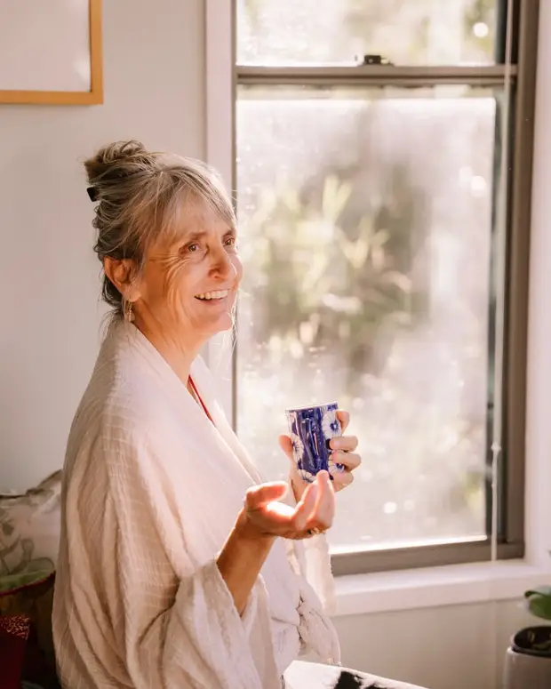 An older woman smiling by a sunlit window with a cup of tea