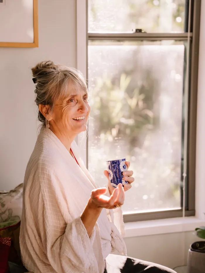 An older woman smiling by a sunlit window with a cup of tea